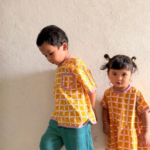 May include: A yellow and white grid-patterned short-sleeved button-down shirt with a purple trim and a chest pocket, paired with teal trousers, alongside a yellow and white grid-patterned short-sleeved dress with a purple trim and a round neckline.