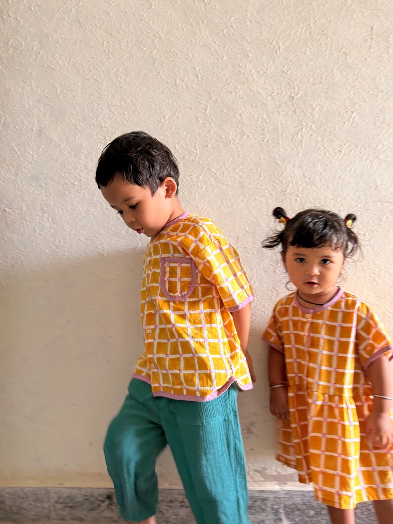 May include: A yellow and white grid-patterned short-sleeved button-down shirt with a purple trim and a chest pocket, paired with teal trousers, alongside a yellow and white grid-patterned short-sleeved dress with a purple trim and a round neckline.
