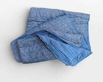 May include: A blue quilted blanket with a geometric pattern. The blanket has a diamond-shaped design in shades of blue and white. The blanket is folded, revealing a solid blue underside.