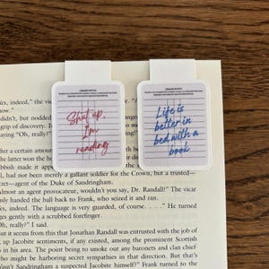 Peut inclure: Deux marque-pages rectangulaires avec des onglets blancs. L'un dit "Shut up. I'm reading" à l'encre rouge, l'autre dit "Life is better in bed with a book" à l'encre bleue. Les deux ont un design de papier ligné.