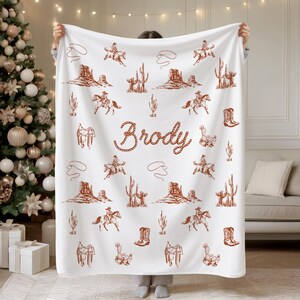Peut inclure: Couverture blanche avec un motif marron sur le th&egrave;me des cow-boys. Le motif comprend des cow-boys &agrave; cheval, des cactus, des bottes, des selles et le nom "Brody" en police de style corde. La couverture est tenue devant un sapin de No&euml;l.