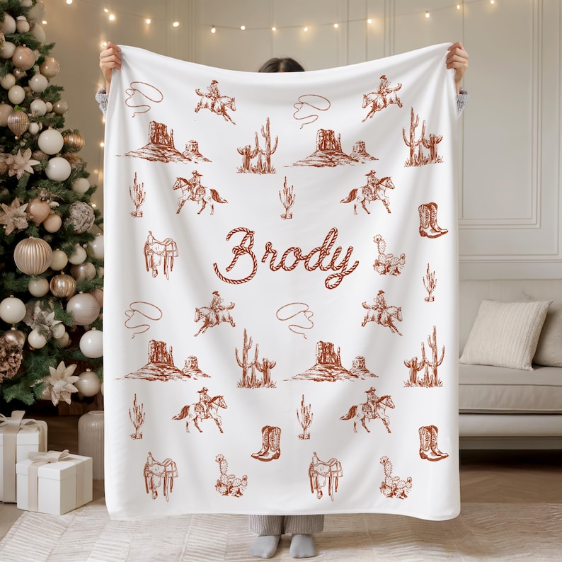Peut inclure: Couverture blanche avec un motif marron sur le th&egrave;me des cow-boys. Le motif comprend des cow-boys &agrave; cheval, des cactus, des bottes, des selles et le nom "Brody" en police de style corde. La couverture est tenue devant un sapin de No&euml;l.