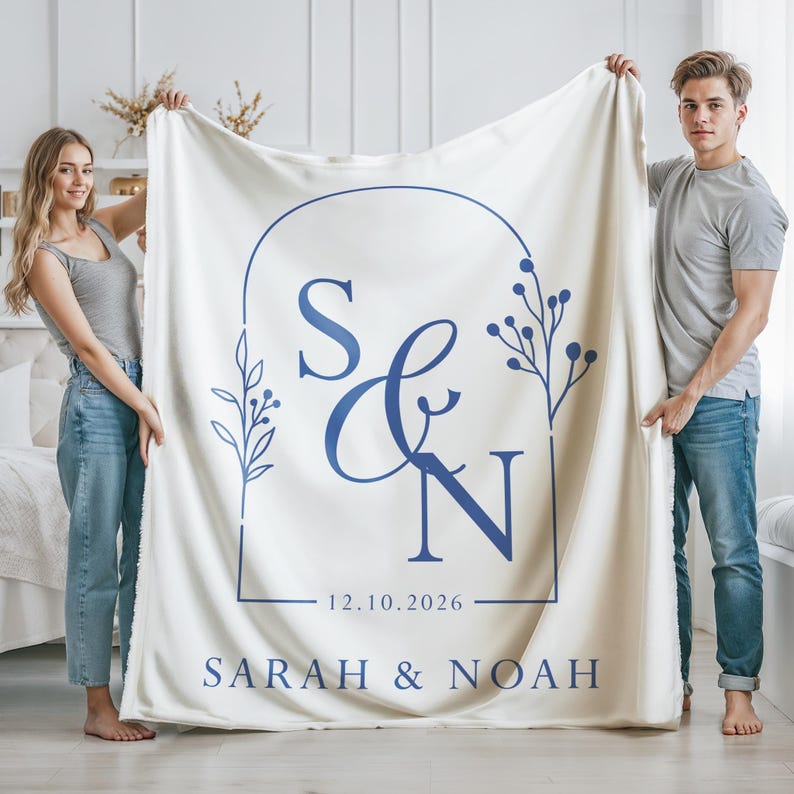 May include: Cream-colored blanket with a blue design featuring the initials "S & N", the date "12.10.2026", and the names "SARAH & NOAH". The blanket is held up by two people.