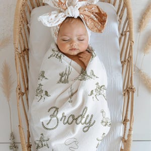 Peut inclure: Un b&eacute;b&eacute; envelopp&eacute; dans une couverture blanche orn&eacute;e d'un motif de cow-boy, avec des cactus et des cow-boys &agrave; cheval. La couverture est personnalis&eacute;e avec le nom "Brody" dans une police de style corde. Le b&eacute;b&eacute; porte un bandeau floral et repose dans un berceau en osier.