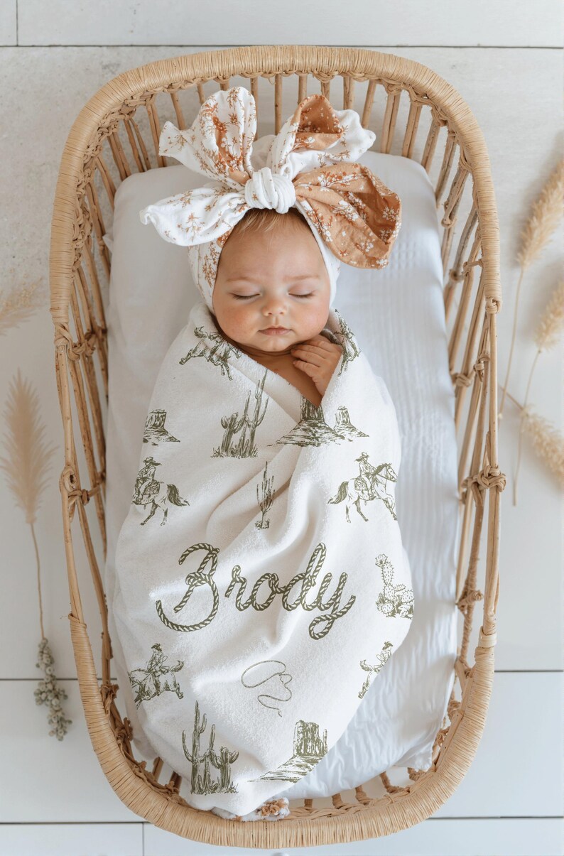 Peut inclure: Un b&eacute;b&eacute; envelopp&eacute; dans une couverture blanche orn&eacute;e d'un motif de cow-boy, avec des cactus et des cow-boys &agrave; cheval. La couverture est personnalis&eacute;e avec le nom "Brody" dans une police de style corde. Le b&eacute;b&eacute; porte un bandeau floral et repose dans un berceau en osier.