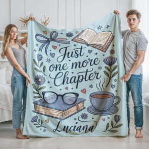 May include: A light blue blanket with the text "Just one more Chapter" and the name "Luciana". The blanket features illustrations of a book, glasses, flowers, a cup of coffee, and hearts. The blanket is held up by two people.