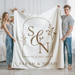 May include: Cream-colored blanket with a brown arched design featuring the initials "S & N", the date "12.10.2026", and the names "SARAH & NOAH". The blanket is held up by two people. The design includes floral elements.