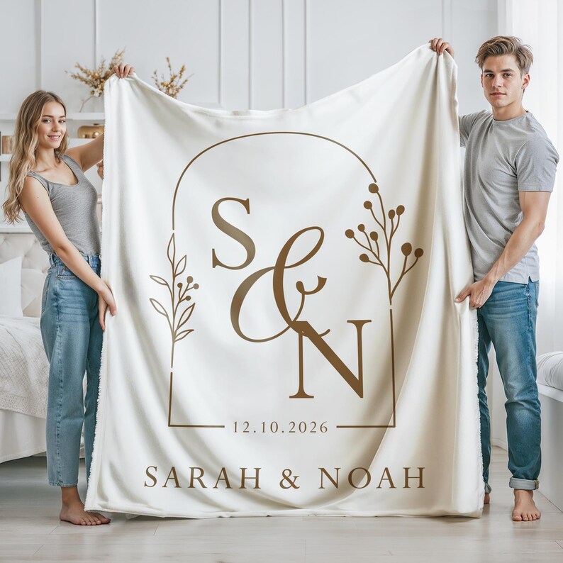 May include: Cream-colored blanket with a brown arched design featuring the initials "S & N", the date "12.10.2026", and the names "SARAH & NOAH". The blanket is held up by two people. The design includes floral elements.