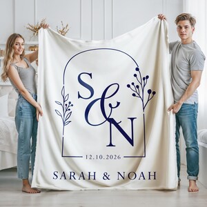 May include: Cream-colored blanket with a navy blue monogram design. The design includes the letters "S & N" within a rectangular frame, with the date "12.10.2026" and the names "SARAH & NOAH" below. The blanket is held up by two people.