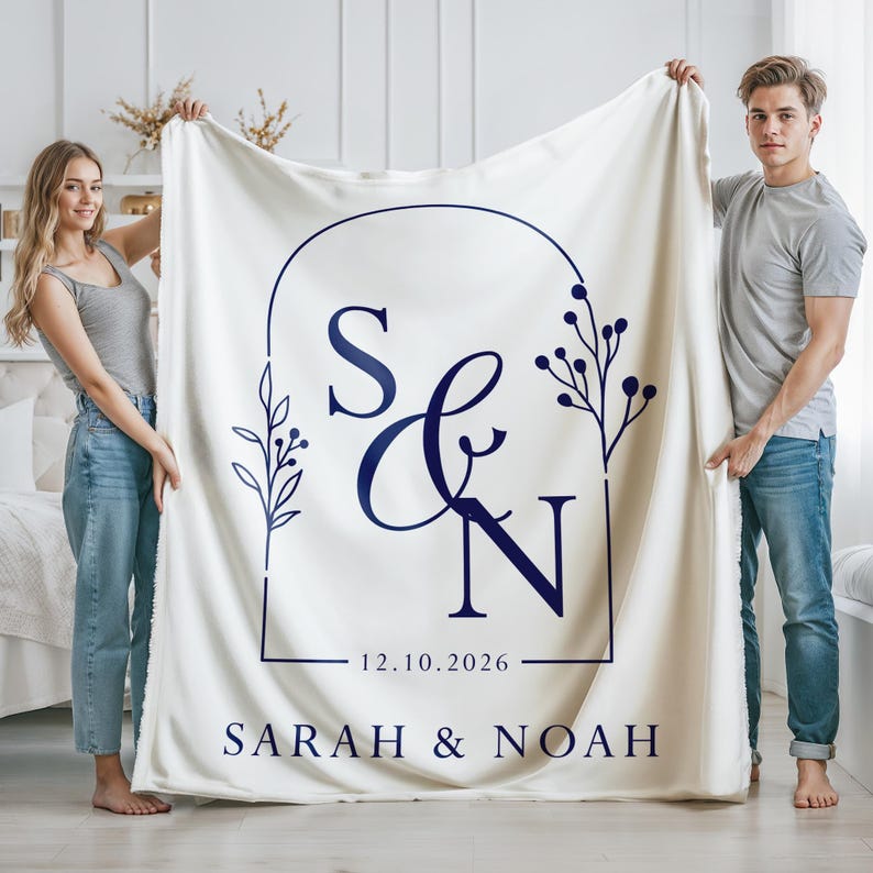 May include: Cream-colored blanket with a navy blue monogram design. The design includes the letters "S & N" within a rectangular frame, with the date "12.10.2026" and the names "SARAH & NOAH" below. The blanket is held up by two people.