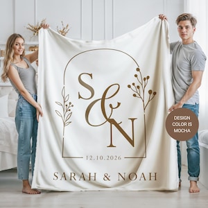 May include: Cream-colored blanket with a mocha-colored design. The design includes the initials "S & N", the date "12.10.2026", and the names "SARAH & NOAH". The blanket is held up by two people. The design color is mocha.