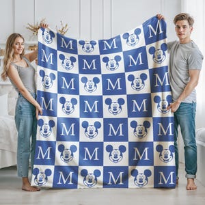 May include: A blue and white checkered blanket featuring Mickey Mouse faces and the letter "M". The blanket is held up by two people. The blanket is a soft, cozy item.