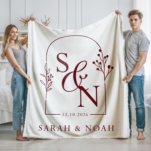 May include: Cream-colored blanket with a burgundy design featuring the initials "S & N", the date "12.10.2026", and the names "SARAH & NOAH". The design is framed by an arch and floral elements.
