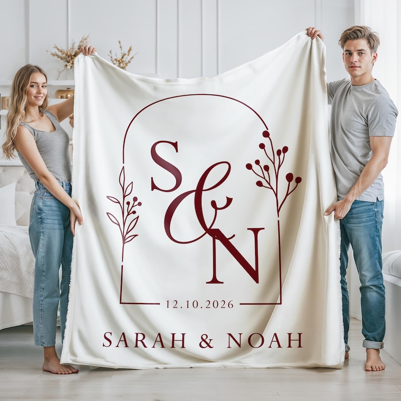 May include: Cream-colored blanket with a burgundy design featuring the initials "S & N", the date "12.10.2026", and the names "SARAH & NOAH". The design is framed by an arch and floral elements.