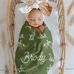 Peut inclure: Un b&eacute;b&eacute; envelopp&eacute; dans une couverture verte sur le th&egrave;me des cow-boys, avec des illustrations blanches de cow-boys, de cactus et le nom "Brody". Le b&eacute;b&eacute; est dans un berceau en osier et porte un bandeau floral.