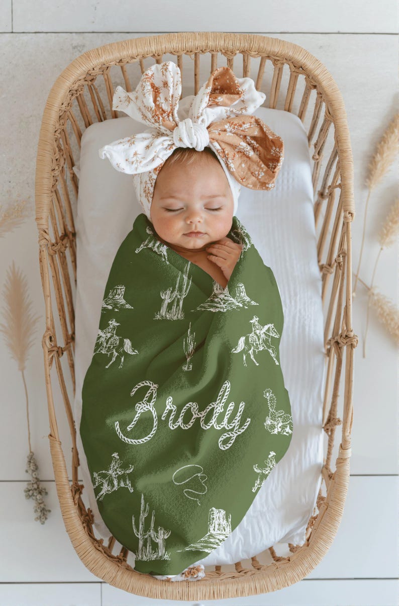 Peut inclure: Un b&eacute;b&eacute; envelopp&eacute; dans une couverture verte sur le th&egrave;me des cow-boys, avec des illustrations blanches de cow-boys, de cactus et le nom "Brody". Le b&eacute;b&eacute; est dans un berceau en osier et porte un bandeau floral.
