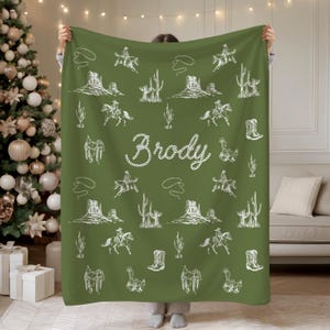 Peut inclure: Une couverture verte orn&eacute;e d'un motif blanc sur le th&egrave;me des cow-boys. Le motif comprend des cow-boys &agrave; cheval, des cactus et des paysages d&eacute;sertiques. Le nom "Brody" est &eacute;crit en blanc.