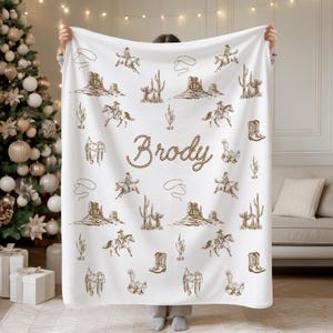 Peut inclure: Plaid blanc avec un motif r&eacute;p&eacute;t&eacute; d'illustrations de cow-boys marron. La couverture pr&eacute;sente des cactus, des chevaux, des selles et le nom "Brody" dans une police de style corde. Le plaid est tenu devant un sapin de No&euml;l et un canap&eacute;.