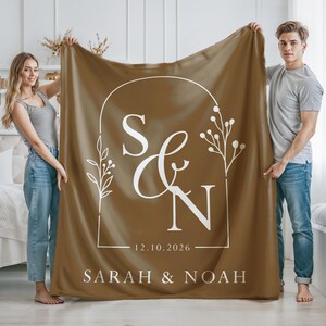 May include: A brown blanket with white text and a decorative floral design. The blanket features the initials "S & N", the date "12.10.2026", and the names "SARAH & NOAH".