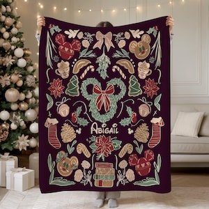 Puede incluir: Una manta de color p&uacute;rpura oscuro con un dise&ntilde;o navide&ntilde;o festivo. La manta est&aacute; adornada con ilustraciones tem&aacute;ticas de vacaciones, que incluyen figuras de pan de jengibre, bastones de caramelo, flores de Nochebuena y una corona. El nombre "Abigail" est&aacute; escrito en cursiva.