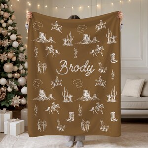 Peut inclure: Une couverture marron avec un motif blanc sur le th&egrave;me des cow-boys, comprenant des cactus, des chevaux et des bottes de cow-boy. Le nom "Brody" est &eacute;crit en cursive. La couverture est tenue par une personne.