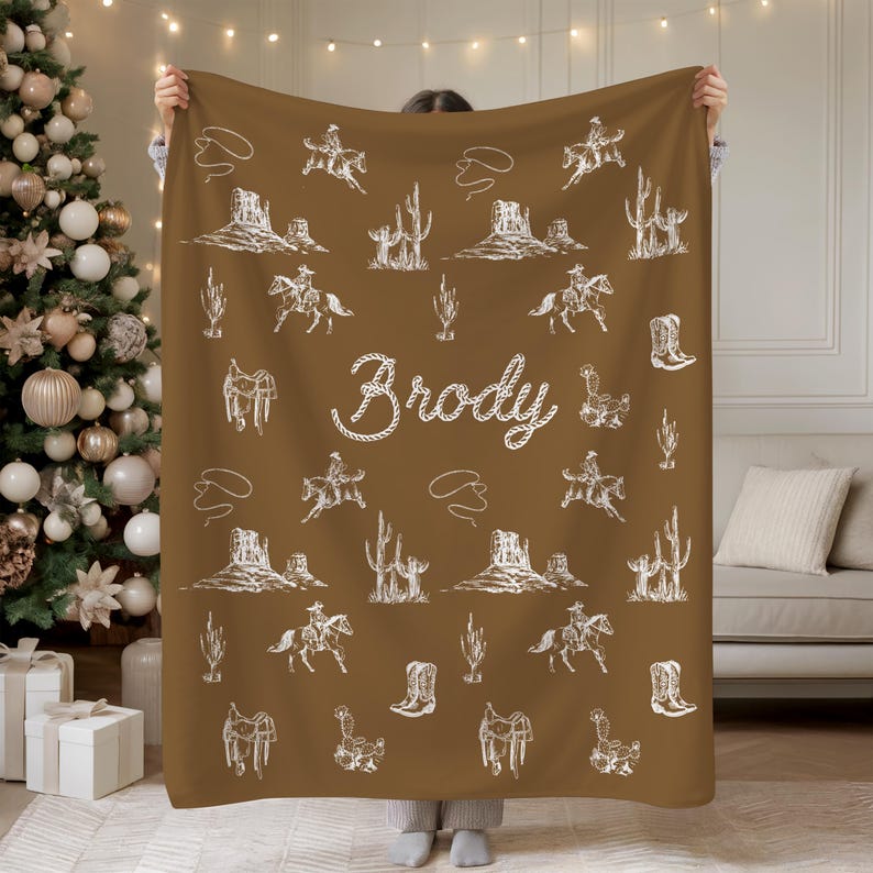 Peut inclure: Une couverture marron avec un motif blanc sur le th&egrave;me des cow-boys, comprenant des cactus, des chevaux et des bottes de cow-boy. Le nom "Brody" est &eacute;crit en cursive. La couverture est tenue par une personne.