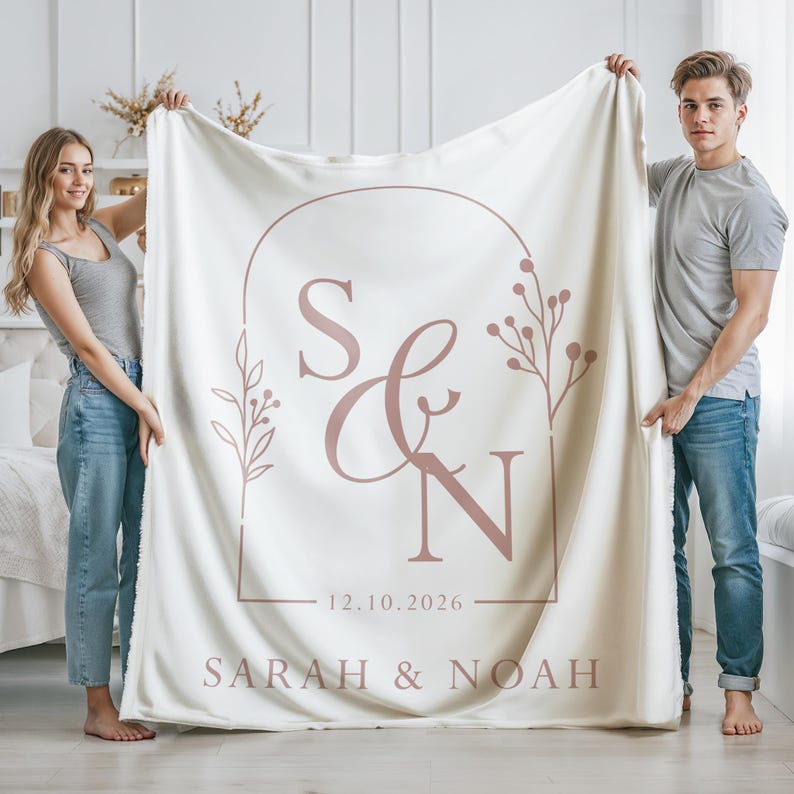 May include: Cream-colored blanket featuring a custom design with the initials "S & N" inside a rectangular arch, with the date "12.10.2026" and the names "SARAH & NOAH" printed below. The design includes floral accents.
