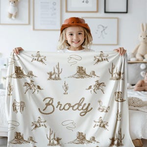 Peut inclure: Une couverture cr&egrave;me avec un motif sur le th&egrave;me du western, repr&eacute;sentant des cow-boys &agrave; cheval, des cactus et des paysages d&eacute;sertiques. Le nom "Brody" est affich&eacute; dans une police de style corde. L'enfant porte un chapeau de cowboy.
