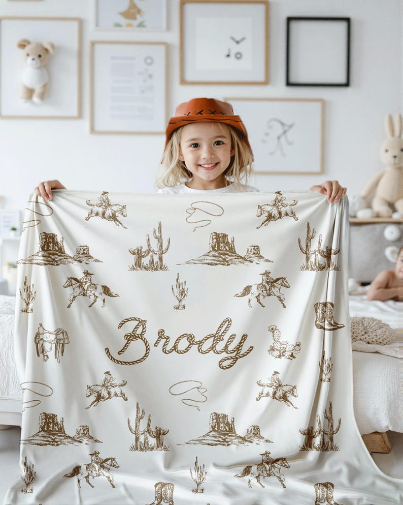 Peut inclure: Une couverture cr&egrave;me avec un motif sur le th&egrave;me du western, repr&eacute;sentant des cow-boys &agrave; cheval, des cactus et des paysages d&eacute;sertiques. Le nom "Brody" est affich&eacute; dans une police de style corde. L'enfant porte un chapeau de cowboy.