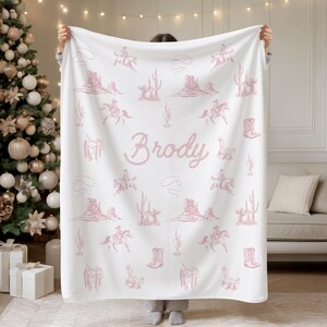 Peut inclure: Une couverture blanche orn&eacute;e d'un motif rose sur le th&egrave;me des cow-boys. Le motif comprend des cow-boys &agrave; cheval, des cactus et des paysages d&eacute;sertiques. Le nom "Brody" est &eacute;crit dans une police de style corde. La couverture est tenue devant un sapin de No&euml;l et un canap&eacute;.