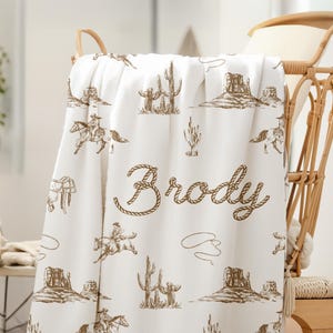 Peut inclure: Couverture de b&eacute;b&eacute; blanche avec un motif marron sur le th&egrave;me des cow-boys. Le motif comprend des illustrations de cow-boys &agrave; cheval, de cactus et de paysages d&eacute;sertiques. Le nom "Brody" est &eacute;crit dans une police de style corde.