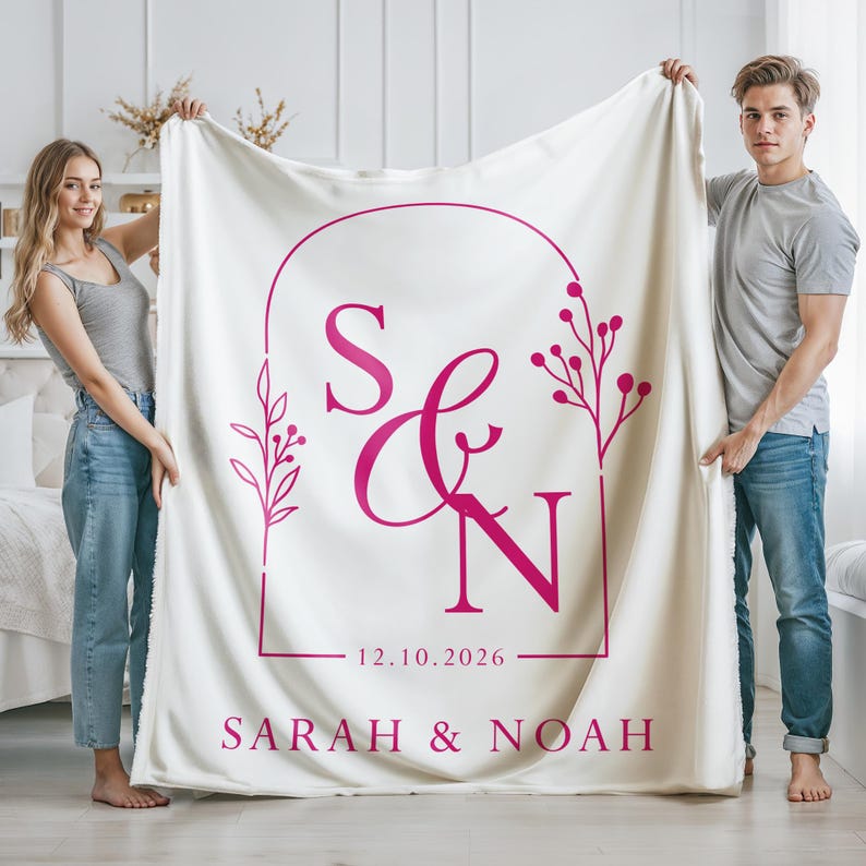May include: A cream-colored blanket with a magenta design featuring the initials "S & N", the date "12.10.2026", and the names "SARAH & NOAH". The design is framed by an arch and floral elements.