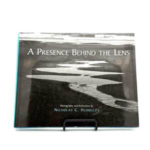 May include: A book titled "A Presence Behind the Lens" with a black and white photograph of a shoreline. The book has a teal spine and is displayed on a black metal stand. The author's name is Nicholas C. Hlobeczy.