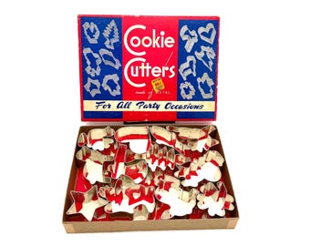 May include: A vintage box of metal cookie cutters. The box is red and blue with the words "Cookie Cutters" and "For All Party Occasions". The cookie cutters are various shapes, including stars and animals, and are displayed in a cardboard box.