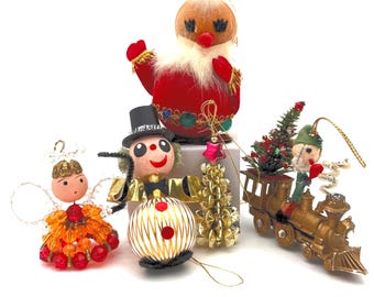 May include: A collection of five vintage Christmas ornaments including a red velvet Santa, a beaded angel, a clown with a top hat, a gold metallic tree, and a small figure riding a gold train with a miniature green pine tree attached.