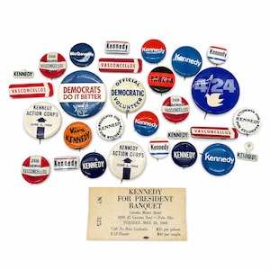 May include: A collection of vintage political campaign buttons and pins featuring various Kennedy, Vasconcellos, and Democratic slogans, alongside a rectangular paper ticket for a 1968 Kennedy for President banquet at the Cabaña Motor Hotel.