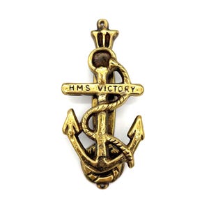 May include: A brass-colored anchor-shaped door knocker with the text "HMS VICTORY" on a horizontal bar. The anchor has a rope wrapped around it and a crown at the top. The knocker is designed to be mounted on a door.