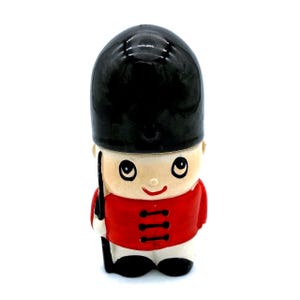 Könnte beinhalten: Keramikfigur eines britischen Royal Guard, mit schwarzem Bärenfellhut und roter Tunika mit schwarzen Knöpfen. Die Figur hat ein lächelndes Gesicht und hält ein schwarzes Gewehr. Der Sockel ist weiß.