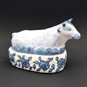 Puede incluir: Una mantequera de cerámica con forma de vaca. La vaca blanca tiene detalles azules y se asienta sobre una base adornada con diseños florales azules. Este plato es para mantequilla y añade un elemento decorativo a la cocina.