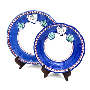 May include: Two blue and white ceramic plates with a bird and leaf design. The plates have a blue border with red dots and a red inner ring. The plates are displayed on brown wooden stands.
