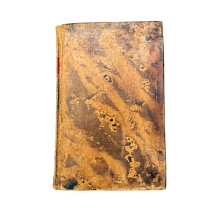 May include: A vintage book with a worn, textured cover. The cover is primarily brown with darker brown and black spots, and a reddish spine. The edges show signs of wear, suggesting age and use. The book is set against a white background.