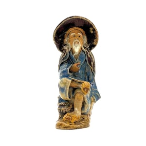 Op de afbeelding: Een keramieken beeldje van een oude man die zit, met een breedgerande hoed en blauwe gewaden. De figuur heeft een lange witte baard en houdt een voorwerp vast. Het beeldje is geschilderd in tinten bruin, blauw en beige.