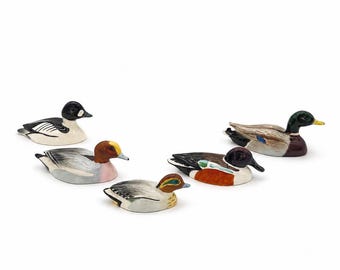 May include: A collection of six ceramic duck figurines in various colors and patterns. The ducks are in a resting position, with detailed plumage and realistic features. The color palette includes shades of brown, white, black, and green.