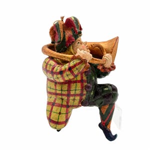 May include: A jester figurine wearing a red, yellow, and green plaid jacket and a matching multi-pointed hat. The figure is playing a large, curved, gold-colored horn. The jester has dark green pants with a diamond pattern and pointed shoes.