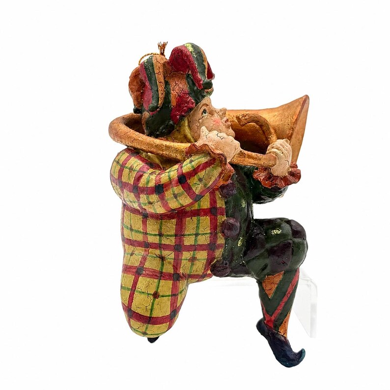 May include: A jester figurine wearing a red, yellow, and green plaid jacket and a matching multi-pointed hat. The figure is playing a large, curved, gold-colored horn. The jester has dark green pants with a diamond pattern and pointed shoes.