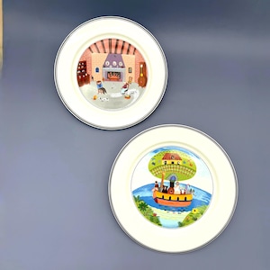 Könnte beinhalten: Zwei dekorative Teller mit cremefarbenem Rand und schwarzem Zierstreifen. Einer zeigt eine Kamin-Szene, der andere ein farbenfrohes Boot mit Passagieren. Die Teller sind wahrscheinlich zur Dekoration gedacht.