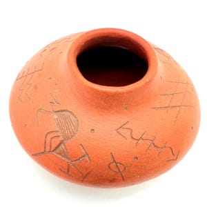 Op de afbeelding: Een terracotta-kleurige, ronde aardewerken vaas met een brede opening. Het oppervlak is versierd met geëtste, abstracte ontwerpen en een gestileerde menselijke figuur. De vaas heeft een gladde, ongeglazuurde afwerking, die de handgemaakte kwaliteit laat zien.