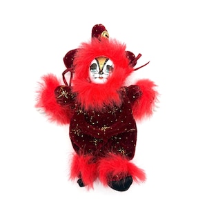 Puede incluir: Muñeca vintage vestida con un atuendo de terciopelo rojo con detalles de estrellas doradas y adornos de plumas rojas. La muñeca tiene una cara pintada con ojos azules y una expresión caprichosa. El atuendo incluye una capucha y puños a juego.