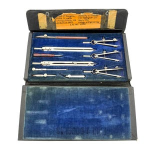 May include: A vintage black leather drafting instrument case with a blue velvet interior containing various metal compasses, dividers, and drawing tools. A yellowed identification label is attached to the inside lid with printed text.