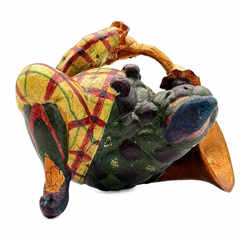 May include: A decorative figurine featuring a rounded, textured body with a dark green and purple diamond pattern. The figure has a yellow and red plaid section, a curved gold-toned handle, and a cracked, weathered paint finish throughout.